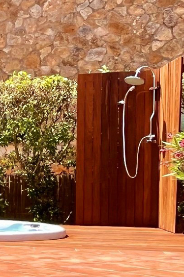 Outdoor shower with a curved handheld shower head mounted on a wooden privacy wall beside a pool on a sunny day.