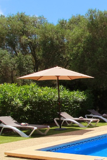 Sunlit poolside scene with white lounge chairs, a large beige parasol, and green hedges surrounding a blue swimming pool.